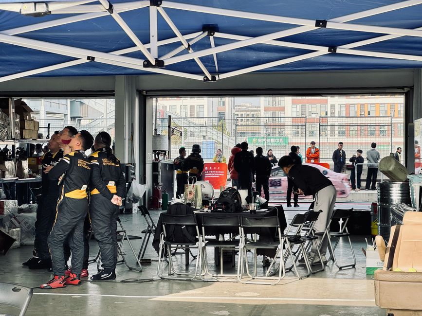A racing team preps in a pit garage while cars zoom past outside, highlighting teamwork and focus.